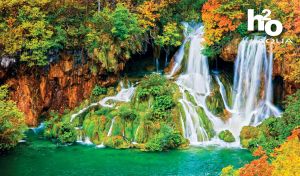 Three-stream waterfall falls from a rocky cliff of green and red trees into the river