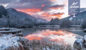 Grey mountains and orange-pink sunset clouds reflected in the winter lake
