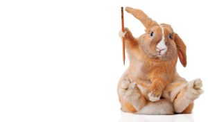 A ginger rabbit draftsman with blue eyes and white nose sits and holds a pencil in a paw